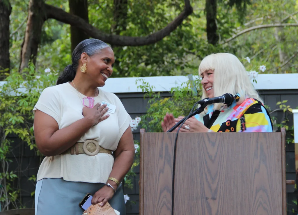 C4DP’s outgoing and incoming executive directors share a warm moment at an outdoor event, speaking at a podium about supporting survivors and promoting healthy, safe relationships.