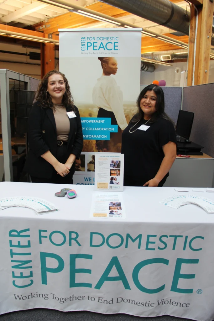 Two C4DP staff members stand behind an indoor outreach table featuring brochures, pledge materials, and a banner promoting empowerment, community collaboration, and transformation.