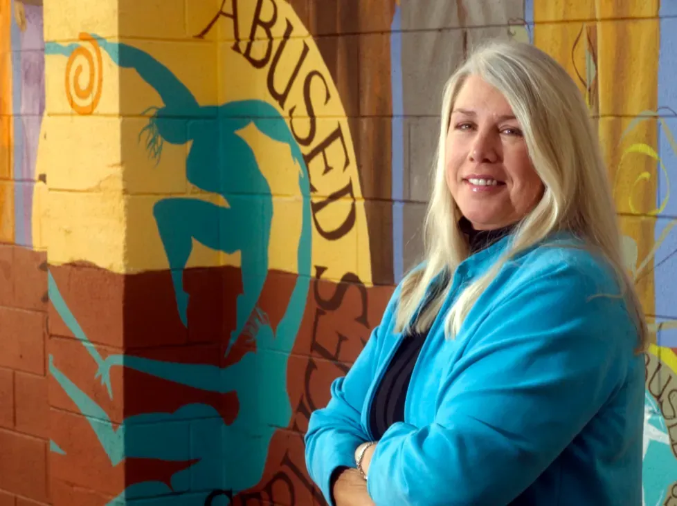 Former Center for Domestic Peace executive director Donna Garske stands smiling in front of a mural at the organization’s Marin County office, honoring her leadership in domestic violence prevention.