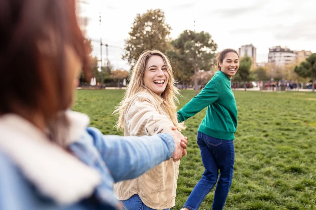 Three young women hold hands in a line while walking through a park, smiling and laughing together on a sunny day.