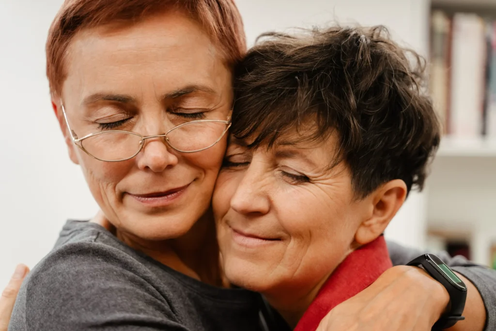 Two older women in a same-sex couple embrace with their eyes closed, sharing a warm and supportive hug.