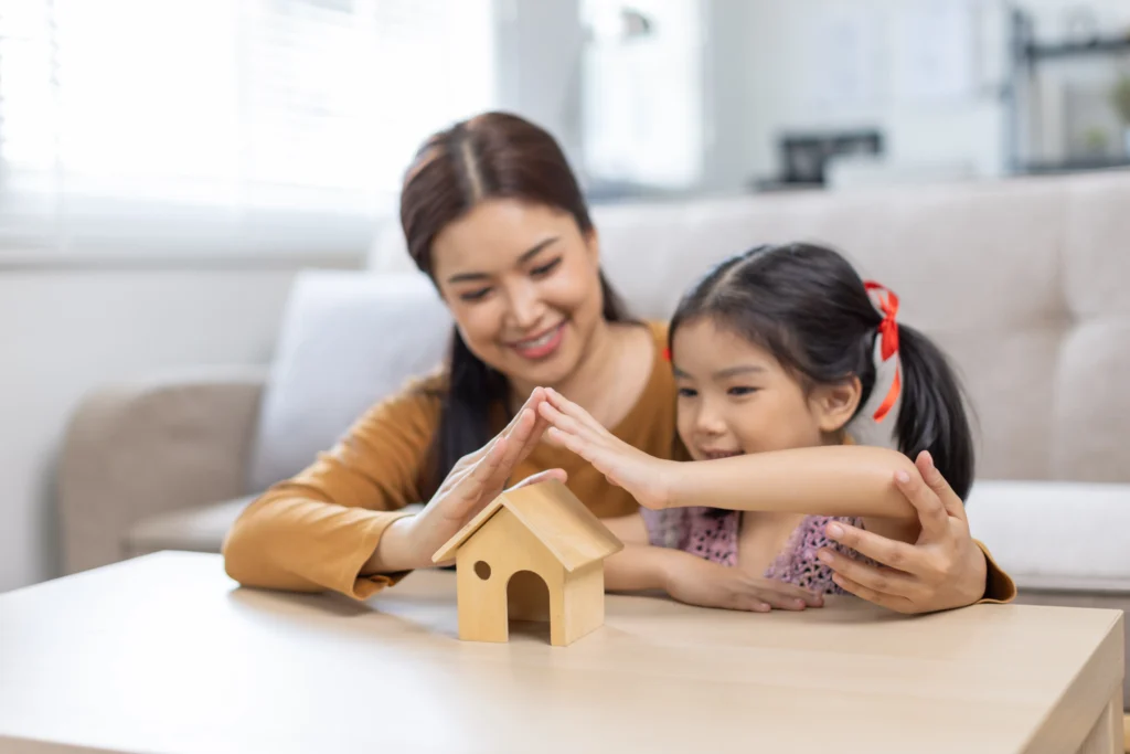 An Asian mother and her young daughter smile as they place their hands together over a small wooden toy house, creating a roof shape to symbolize safety and protection.