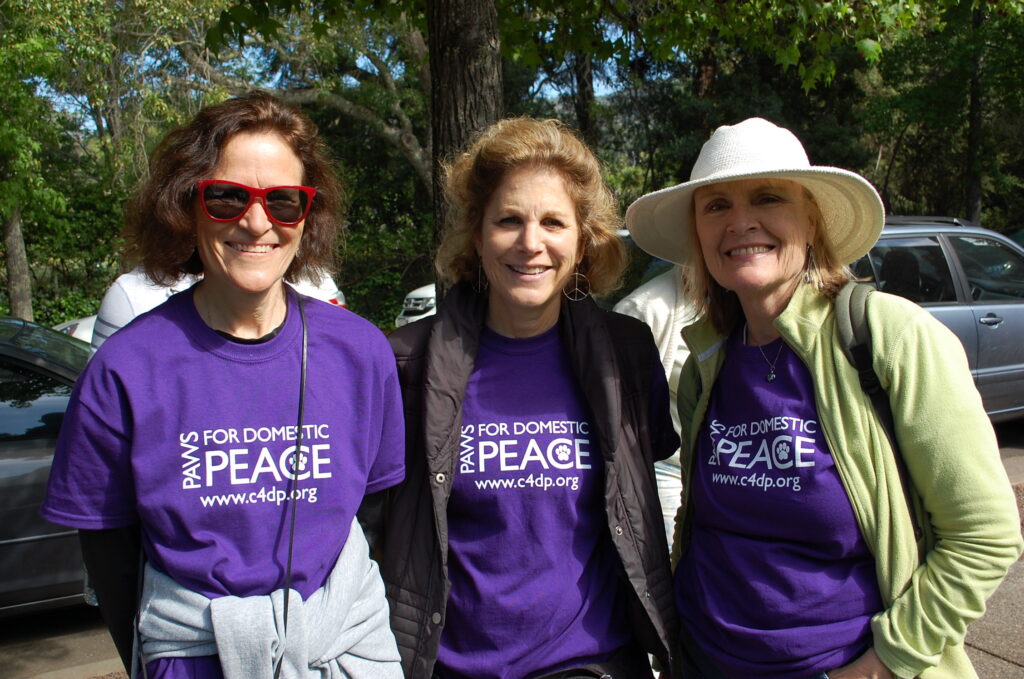 Three C4DP supporters wearing “Paws for Domestic Peace” shirts smile together before a community walk supporting survivors in Marin County.