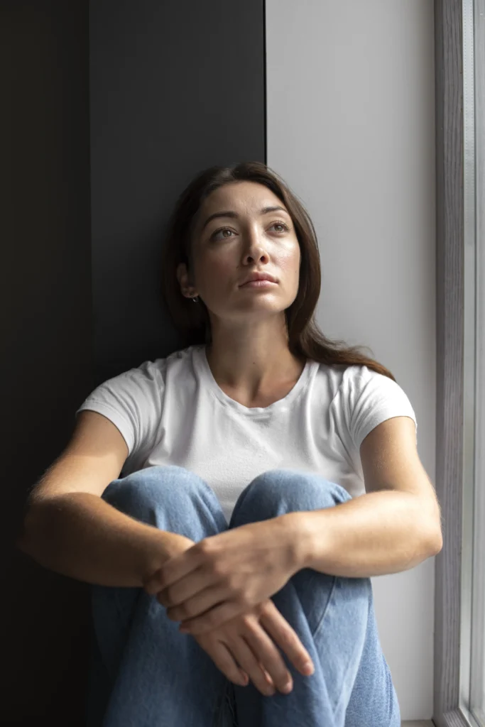 A young woman with sits by a window with her knees pulled close, looking upward with a thoughtful, distant expression as natural light falls across her face.