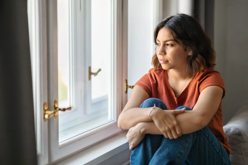 A teenage girl sits by a window with her knees drawn in, looking out with a tense, worried expression, illustrating how domestic violence can affect children and teens.