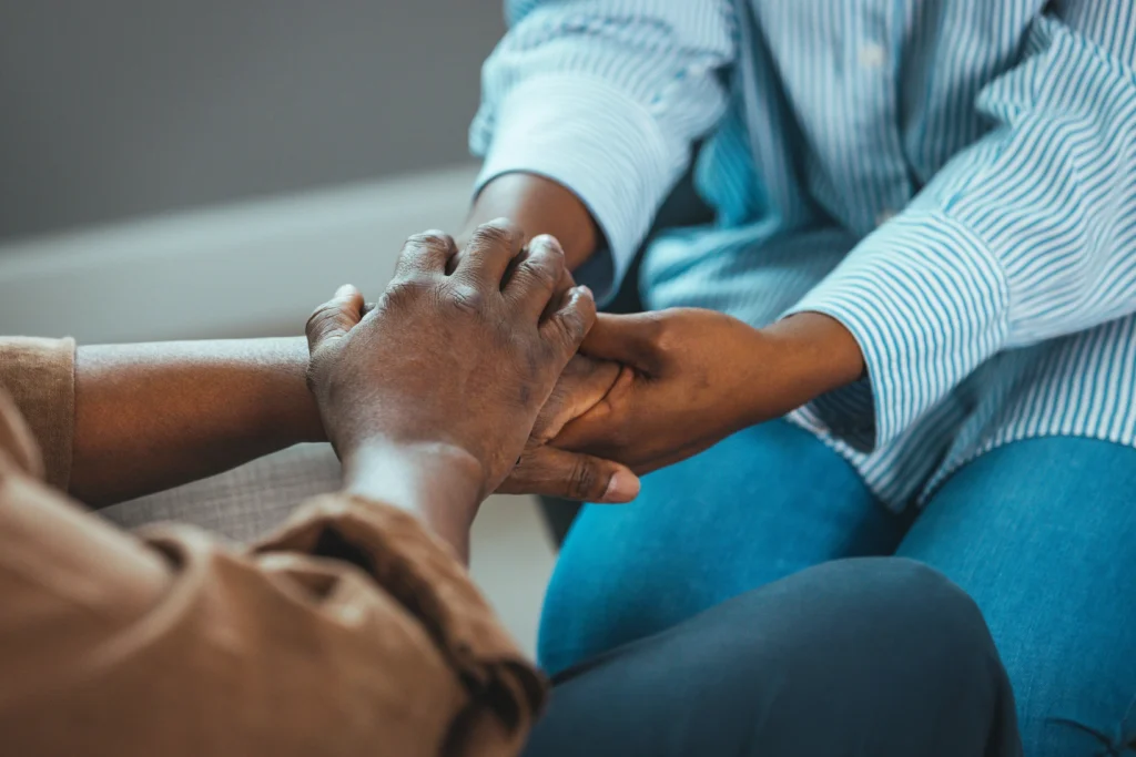 Two Black individuals sit close together as one gently holds the other’s hands in a supportive, comforting gesture.